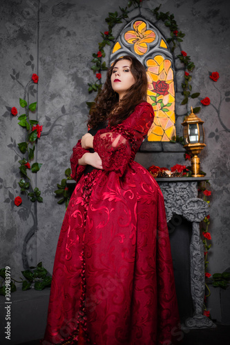 A beautiful girl in a magnificent red dress of the Rococo era stands with a book in her hands against the background of a fireplace, window and flowers.