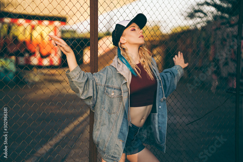 Young attractive woman in jeans jacket, shorts, red top and trucker hat posing over metal fence and graffiti wall over background in a city