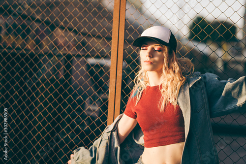 Young attractive woman in jeans jacket, shorts, red top and trucker hat posing over metal fence and graffiti wall over background in a city