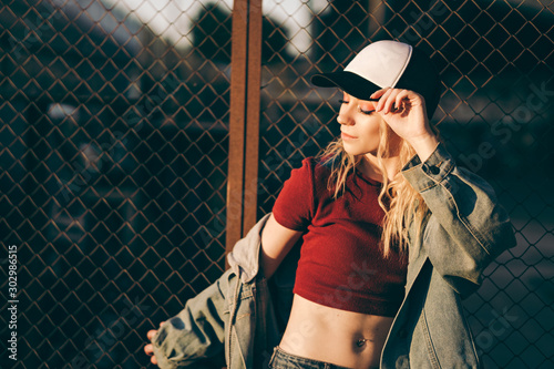 Young attractive woman in jeans jacket, shorts, red top and trucker hat posing over metal fence and graffiti wall over background in a city