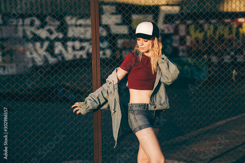 Young attractive woman in jeans jacket, shorts, red top and trucker hat posing over metal fence and graffiti wall over background in a city