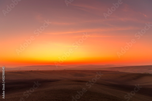 The sun caught during the last moment before setting beyond the horizon with view of a field covered with fog and the farms and fields undulating in the region of South Moravia is also called Tuscany