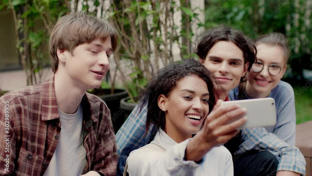 Close up shot of young cheerful multinational students happily taking selfie on cellphone resting in university campus outdoor Close up shot of young cheerful multinational students happily taking selfie on cellphone resting in university campus outdoor