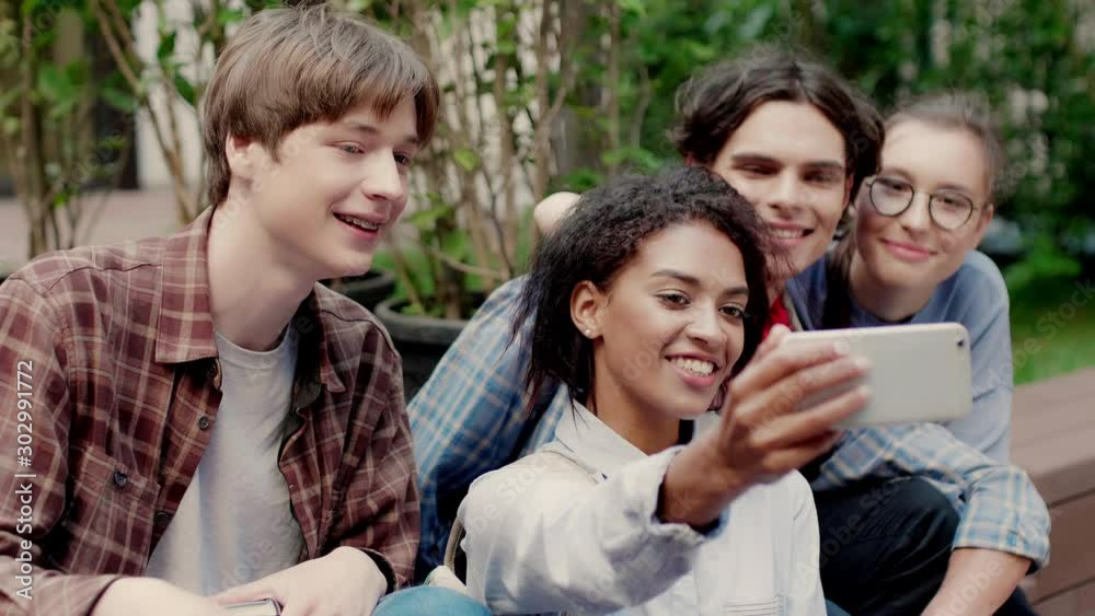 Group of young smiling multinational students joyfully taking selfie on cellphone resting in university campus outdoor