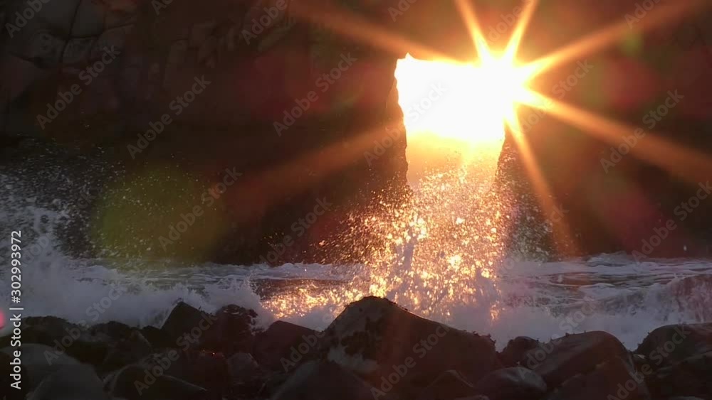 Slow motion waves crash over smooth stone at Pfeiffer Beach as a sunstar forms against the hard rock sea stack in this amazing natural wonder from Western California on the Pacific Ocean coast. 