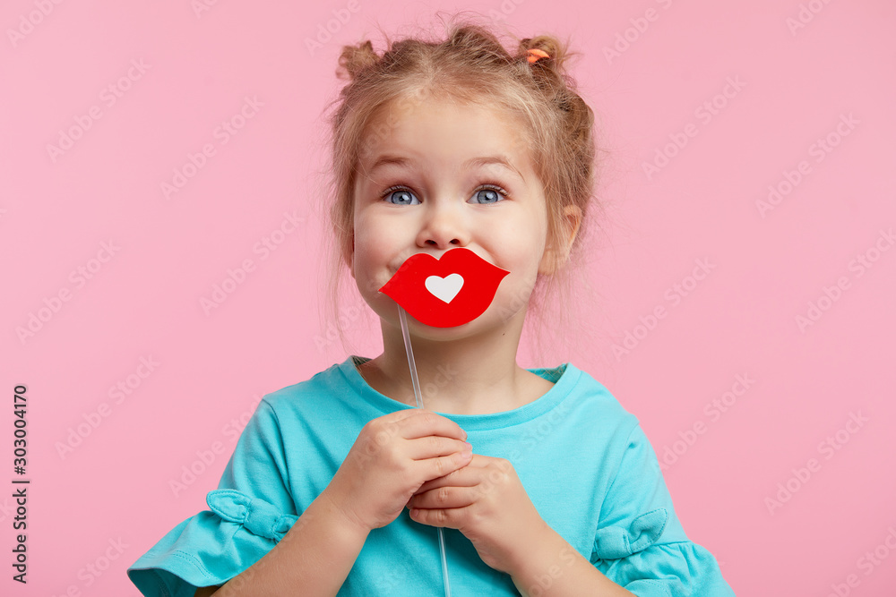Funny smiley girl face on the background of a bright pink wall. Child girl with paper accessories, paper lips on a stick.