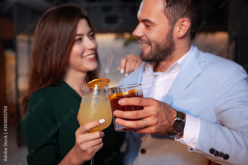 Cropped closeup of a happy loving couple clinking cocktail glasses, celebrating valentines day at the bar. Selective focus on drinks in hands of young couple Cropped closeup of a happy loving couple clinking cocktail glasses, celebrating valentines day at the bar. Selective focus on drinks in hands of young couple