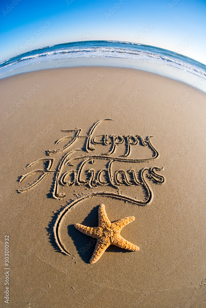 Happy Holidays message written in smooth sand with fisheye effect on starfish and frothy white waves - obrazy, fototapety, plakaty 