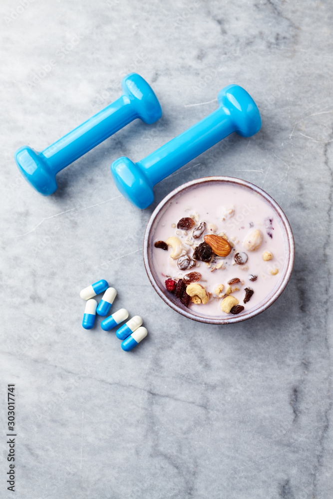 Bowl of granola with yogurt, nuts, raisins, cranberries and chia seeds. Blue dumbbells and sports supplements in background. Concept for a tasty and healthy meal. Stone background. Copy space Bowl of granola with yogurt, nuts, raisins, cranberries and chia seeds. Blue dumbbells and sports supplements in background. Concept for a tasty and healthy meal. Stone background. Copy space