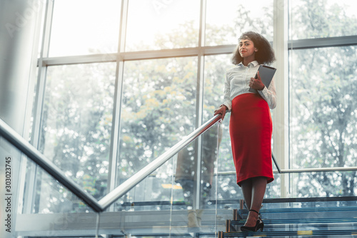 Dazzling African-American businesswoman with a tablet pc is standing on the top of a glass office staircase with chrome railing, a copy space area on the left for an ad text message or your logo
