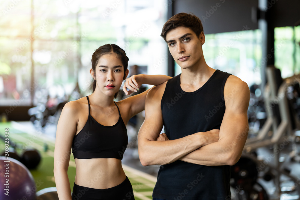 Portrait of young handsome fit Caucasian man and beauty Asian woman standing showing muscle body in gym or fitness club with exercise equipment in background. Working out for health, well being. Portrait of young handsome fit Caucasian man and beauty Asian woman standing showing muscle body in gym or fitness club with exercise equipment in background. Working out for health, well being.
