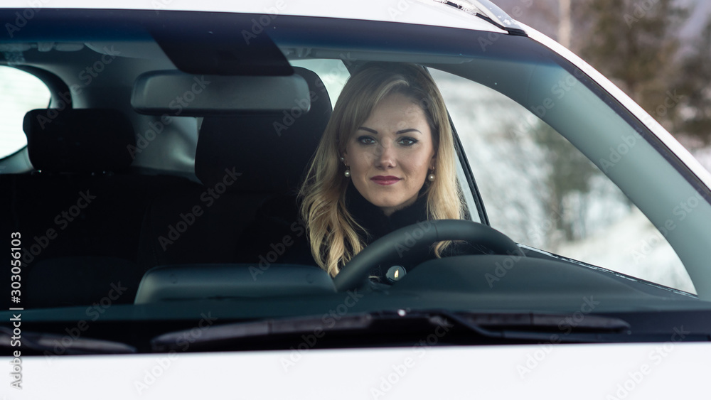 Blonde woman sits at a white car steering and seen through the window.