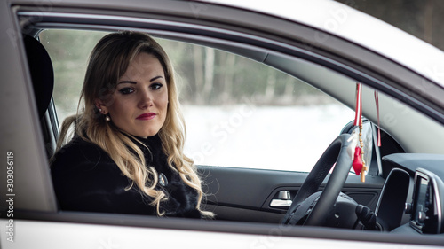 Blonde woman sits at a white car steering wheel and staring through the open window.