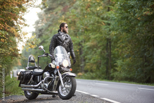 Young bearded tall athletic motorcyclist in dark sunglasses, black leather jacket and gloves standing at shiny modern powerful motorbike on blurred background of hilly asphalt road and green trees.