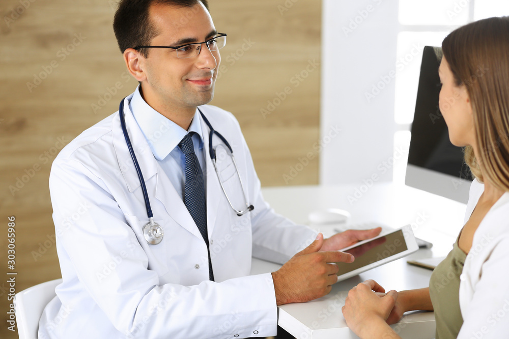 Doctor and patient discussing medical exam results while sitting at the desk in clinic. Male physician using tablet computer for filling up medication history records of young woman. Data in medicine Doctor and patient discussing medical exam results while sitting at the desk in clinic. Male physician using tablet computer for filling up medication history records of young woman. Data in medicine