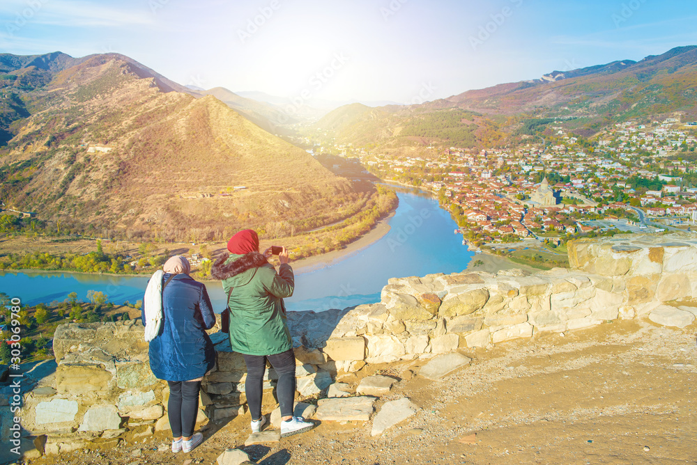 tourists women in winter suit with smartphone take photo of view of Jvari Monastery Georgia with 2 rivers and Svetitskhoveli Cathedral with Mtskheta town in background.