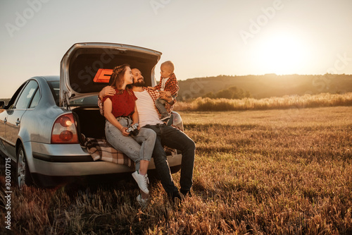 Happy Young Family Mom and Dad with Their Little Son Enjoying Summer Weekend Picnic Sitting on the Trunk of the Car Outside the City in the Field at Sunny Day Sunset, Vacation and Road Trip Concept