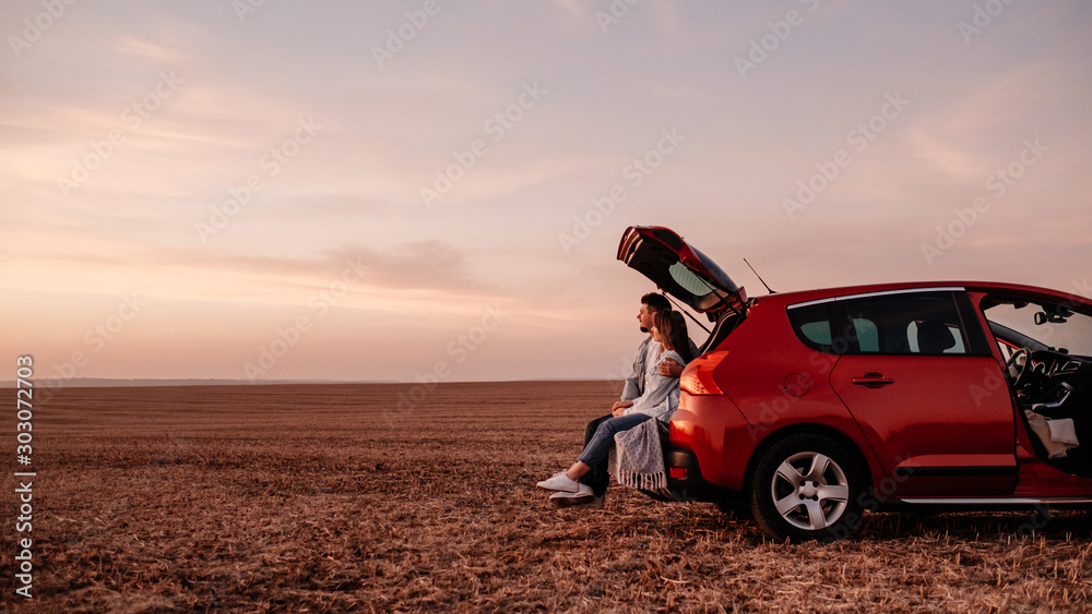 Young Happy Couple Dressed Alike in White Shirt and Jeans Sitting at Their New Car Trunk, Beautiful Sunset on the Field, Vacation and Travel Concept - obrazy, fototapety, plakaty 