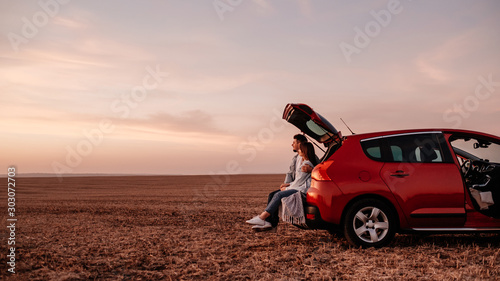 Young Happy Couple Dressed Alike in White Shirt and Jeans Sitting at Their New Car Trunk, Beautiful Sunset on the Field, Vacation and Travel Concept photo