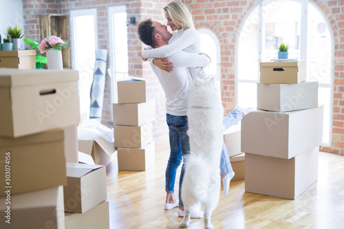 Young beautiful couple with dog hugging at new home around cardboard boxes