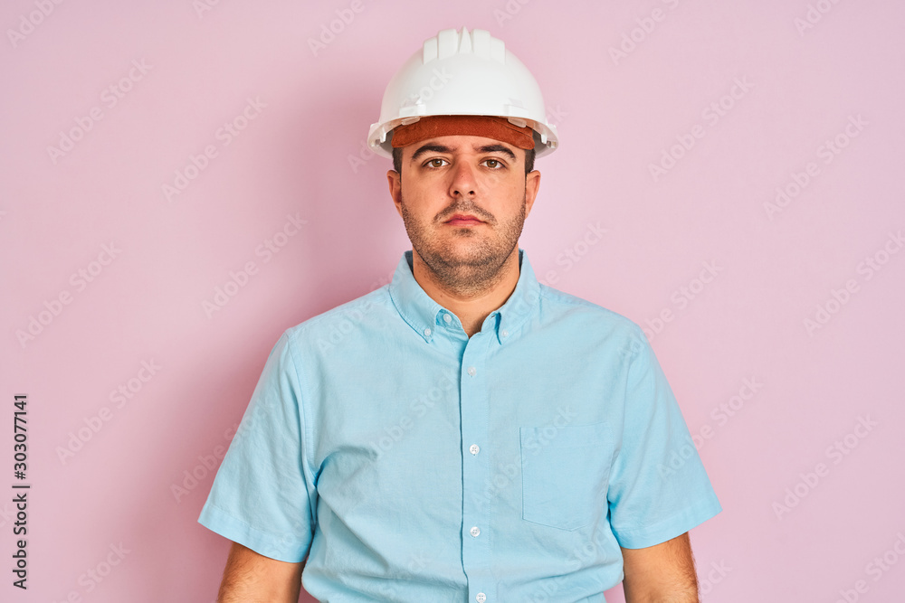 Young architect man wearing security helmet standing over isolated pink background with serious expression on face. Simple and natural looking at the camera.