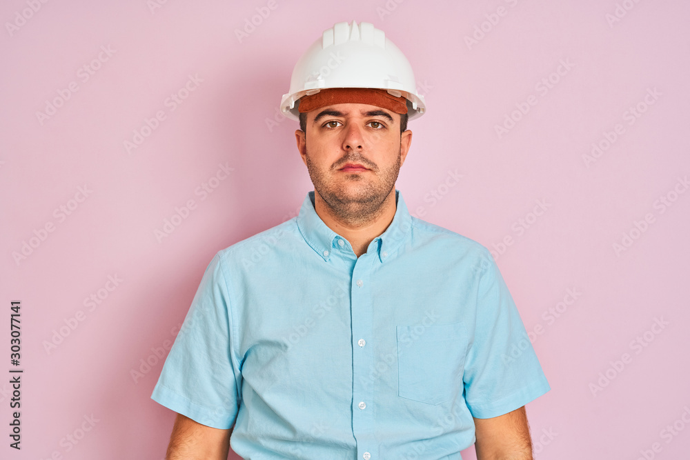 Young architect man wearing security helmet standing over isolated pink background with serious expression on face. Simple and natural looking at the camera. Young architect man wearing security helmet standing over isolated pink background with serious expression on face. Simple and natural looking at the camera.