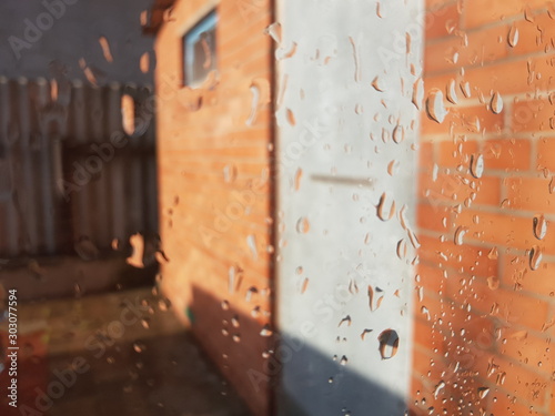 Window in raindrops close-up. Drops of rain on the glass. Orange brick wall with door outdoor as background.