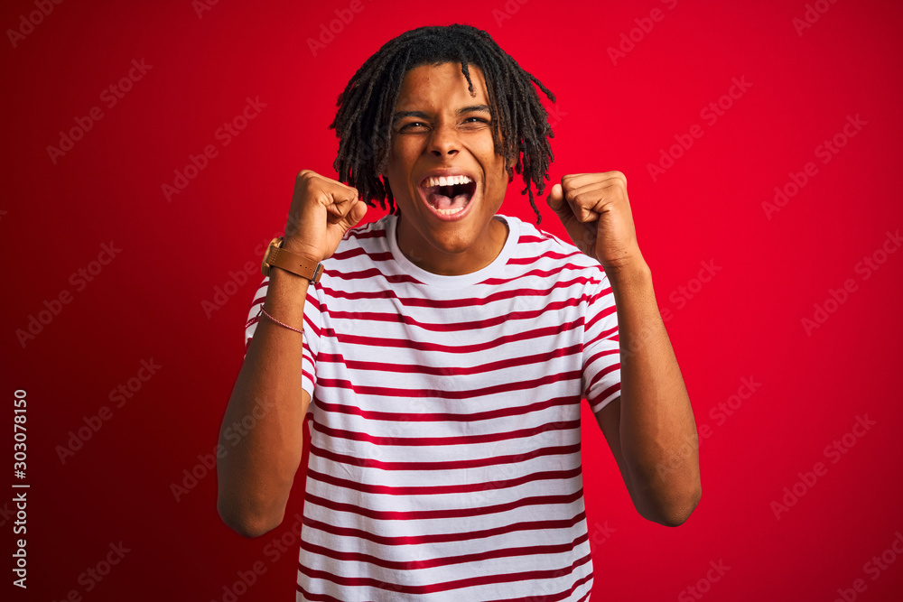 Young afro man with dreadlocks wearing striped t-shirt standing over isolated red background excited for success with arms raised and eyes closed celebrating victory smiling. Winner concept. Young afro man with dreadlocks wearing striped t-shirt standing over isolated red background excited for success with arms raised and eyes closed celebrating victory smiling. Winner concept.
