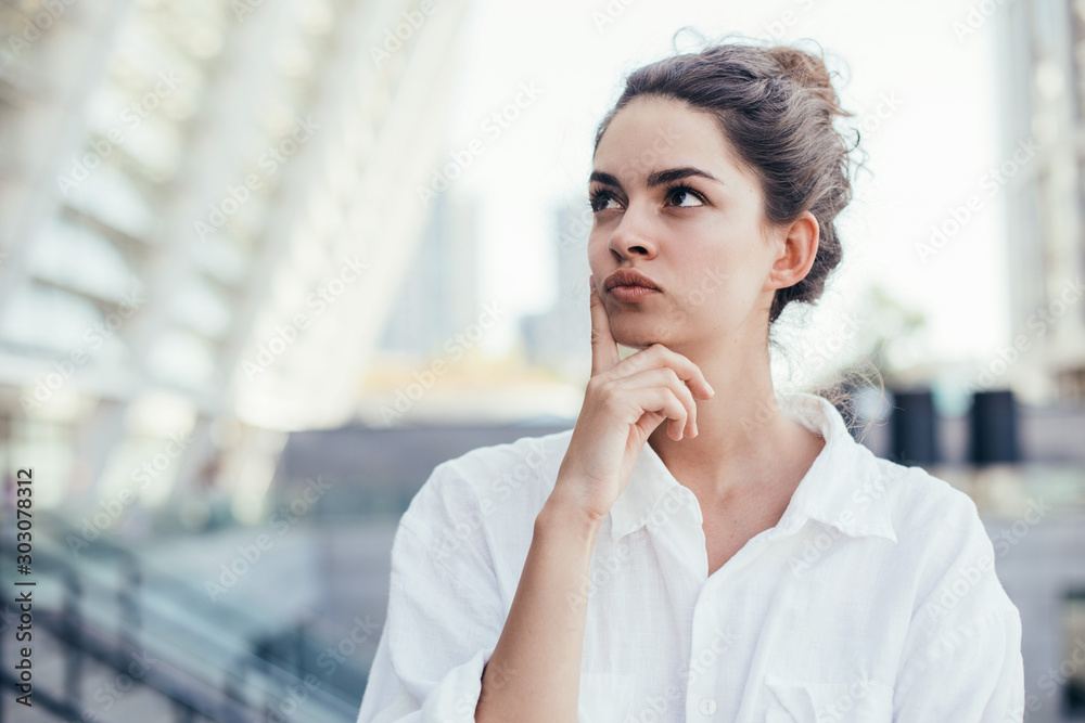 Portrait of calm and thoughtful young woman posing on camera. Stand and look to left. Modern buildings behind. Stand alone outside. Pretty beautiful woman. Portrait of calm and thoughtful young woman posing on camera. Stand and look to left. Modern buildings behind. Stand alone outside. Pretty beautiful woman.