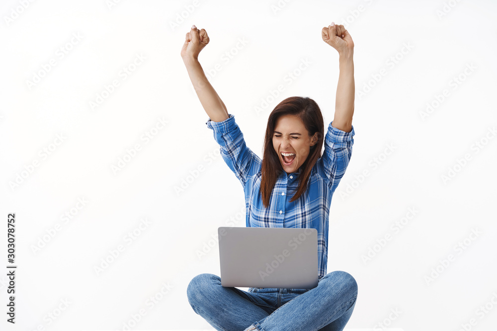 Cheerful ambitious good-looking adult woman express happiness excitement, fist pump, raise hands triumphing sit crossed legs table, hold smartphone laptop, receive excellent news via computer