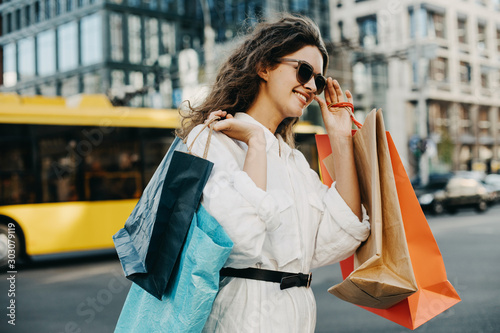 Attractive stylish young woman walking on street and smilw. Wear sunglasses. Shopaholic after shopping. Street style. photo