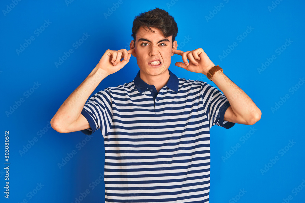Fototapeta premium Teenager boy wearing casual t-shirt standing over blue isolated background covering ears with fingers with annoyed expression for the noise of loud music. Deaf concept.