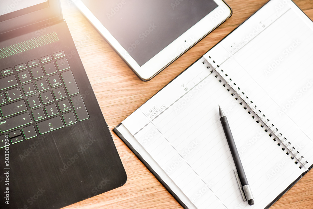 Desk laptop or modern computer with blank screen on rustic table. Spiral notepad placed on wooden desktop with pen and backlight. Business notebook detail concept.