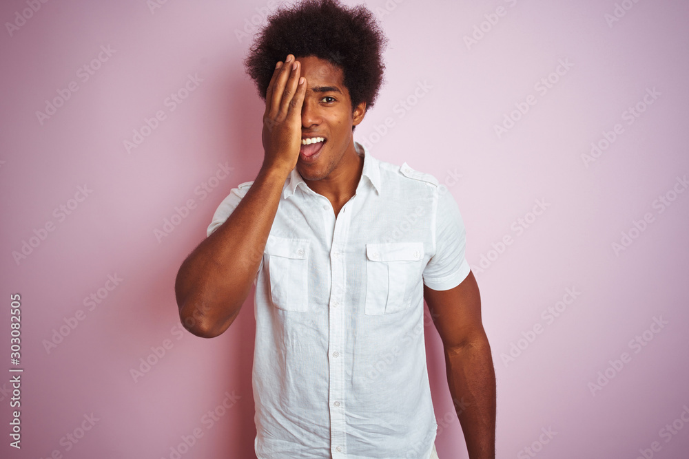 Young american man with afro hair wearing white shirt standing over isolated pink background covering one eye with hand, confident smile on face and surprise emotion. Young american man with afro hair wearing white shirt standing over isolated pink background covering one eye with hand, confident smile on face and surprise emotion.
