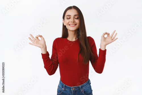 Cheerful, relaxed and patient good-looking brunette girl in red sweater, smiling delighted, close eyes, hold hands sideways in yoga, meditation zen gesture, standing peaceful white background
