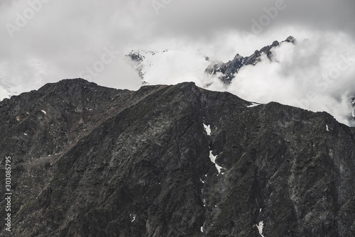 Atmospheric minimalist alpine landscape with big rocky mountain wall and snowbound peak in low clouds. Huge mountains with massive glacier. Craggy wall with snow. Majestic scenery on high altitude.