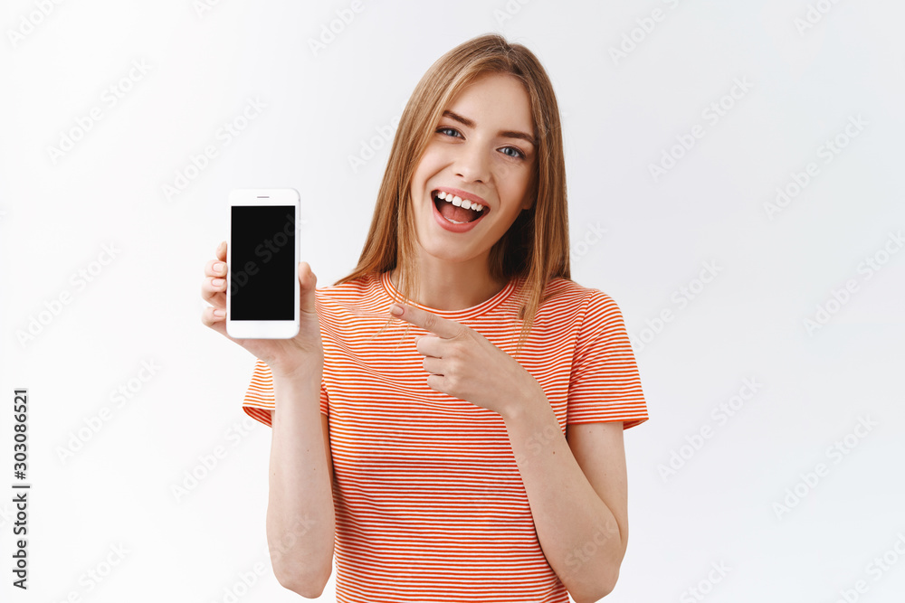 Fototapeta premium Pretty, cheerful young caucasian woman in striped t-shirt holding smartphone, pointing black mobile phone display and smiling as discuss new app, recommend follow her social media, white background