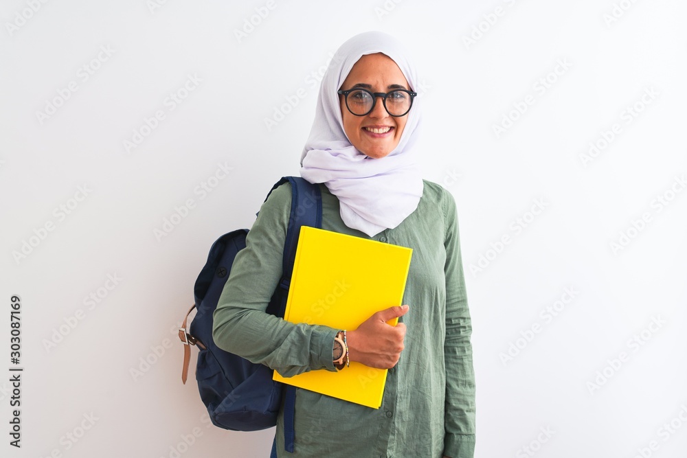 Young Arab student woman wearing hijab and backpack holding a book over isolated background with a happy face standing and smiling with a confident smile showing teeth Young Arab student woman wearing hijab and backpack holding a book over isolated background with a happy face standing and smiling with a confident smile showing teeth