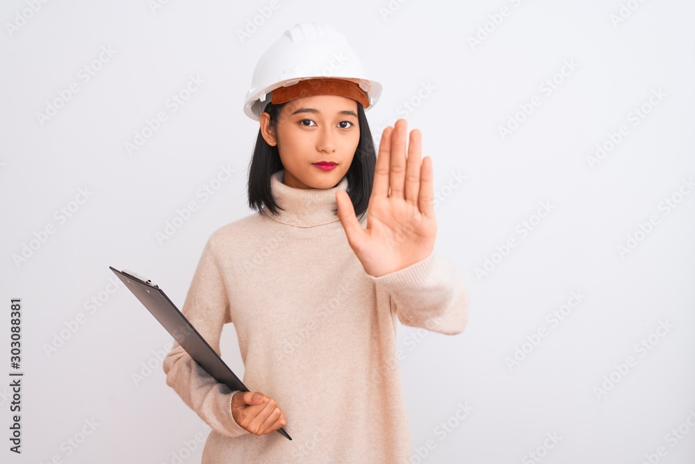 Chinese architect woman wearing helmet holding clipboard over isolated white background with open hand doing stop sign with serious and confident expression, defense gesture Chinese architect woman wearing helmet holding clipboard over isolated white background with open hand doing stop sign with serious and confident expression, defense gesture
