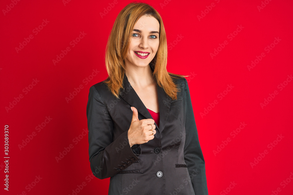 Young caucasian business woman wearing a suit over isolated red background doing happy thumbs up gesture with hand. Approving expression looking at the camera with showing success. Young caucasian business woman wearing a suit over isolated red background doing happy thumbs up gesture with hand. Approving expression looking at the camera with showing success.