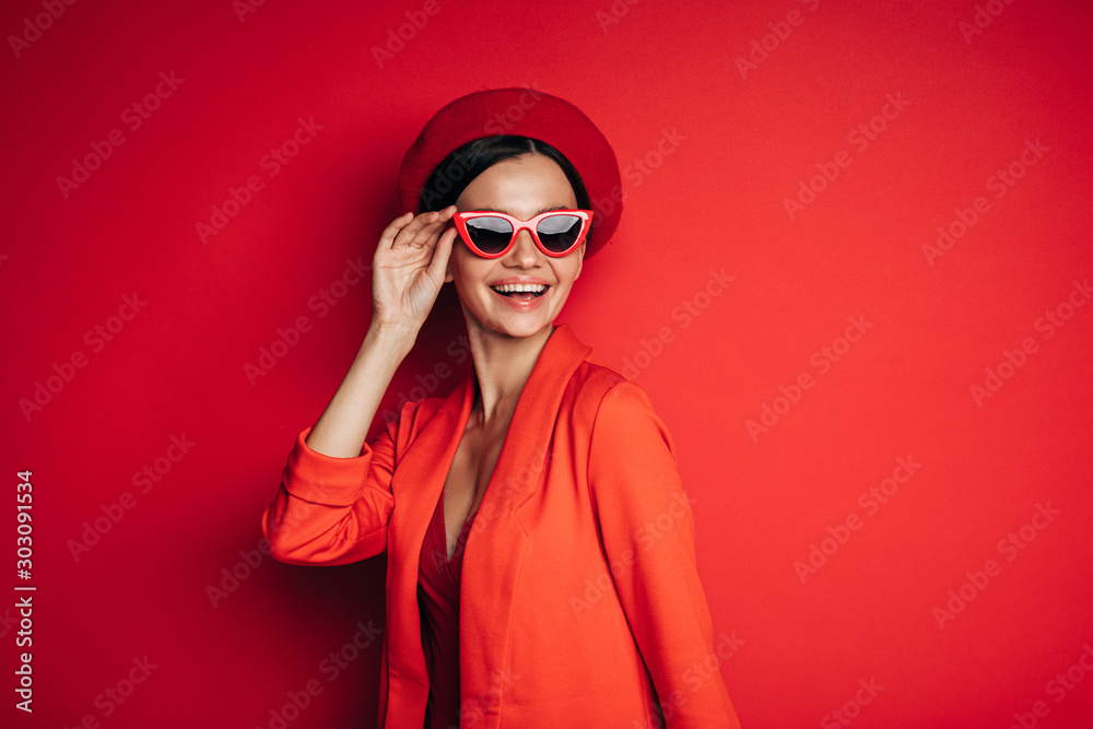 Side view of cheerful happy young woman smiling on set. Wear red beret, jacket. Fashionable model touch sunglasses with hand. Isolated over red background.