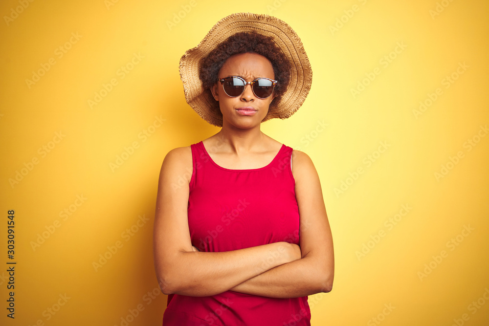 African american woman wearing summer hat and sunglasses over yellow isolated background skeptic and nervous, disapproving expression on face with crossed arms. Negative person. African american woman wearing summer hat and sunglasses over yellow isolated background skeptic and nervous, disapproving expression on face with crossed arms. Negative person.