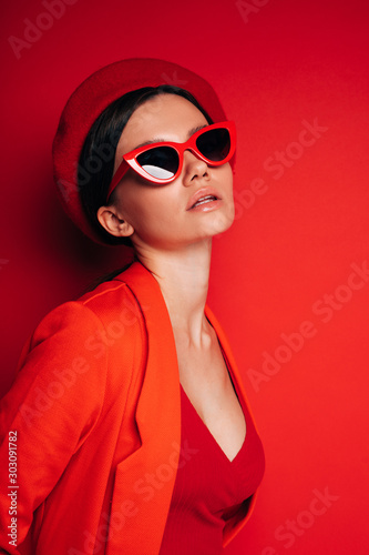Vertical picture of beautiful amazing young woman with dark hair look straight through sunglasses and pose. Wear red clothes and beret. Calm peaceful model. Isolated over red background.
