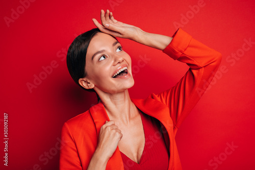 Happy cheerful emotional young woman look up to right and smile. Hold one hand on firehead. Rejoice. Wear stylish red casual clothes. Amazing model. Isolated over red background. photo