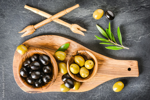 Fresh green and black olives on dark stone table or board. Olive leaves, wooden pickers on black blackground.