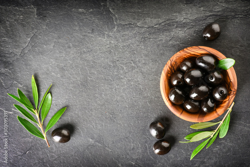 Fresh olives in wooden bowl on dark stone table. Black and green olive with pickers or sticks from top view.