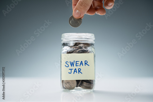 Person Inserting Coin In Swear Jar Over White Desk