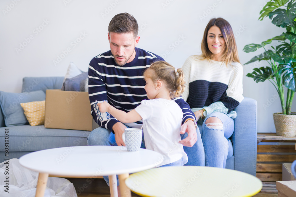 Beautiful family, parents sitting on the sofa drinking coffee looking his kid playing at new home around cardboard boxes