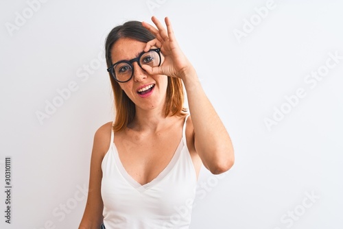 Beautiful redhead woman wearing glasses over isolated background doing ok gesture with hand smiling, eye looking through fingers with happy face.