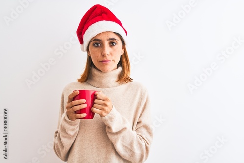 Beautiful redhead woman wearing christmas hat drinking a cup of coffee over isolated background with a confident expression on smart face thinking serious